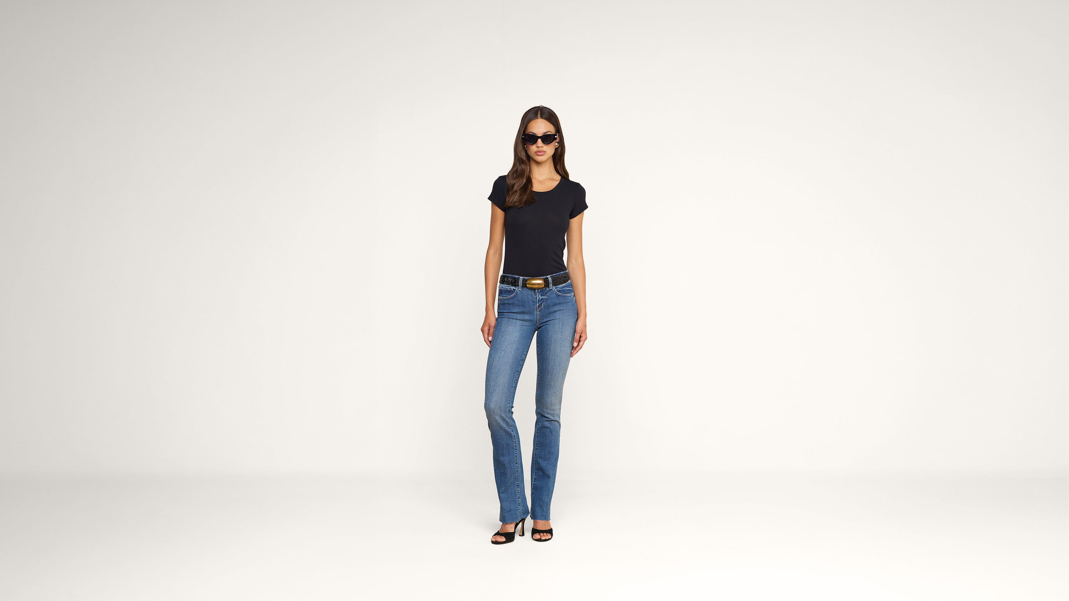 A woman wearing sunglasses, a black t-shirt, blue jeans, and black heeled sandals stands against a plain white background.