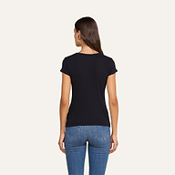 Una donna dai lunghi capelli castani, che indossa una maglietta nera a scollo rotondo di Cory Cotton e dei jeans blu, è in piedi di spalle alla fotocamera su uno sfondo beige.