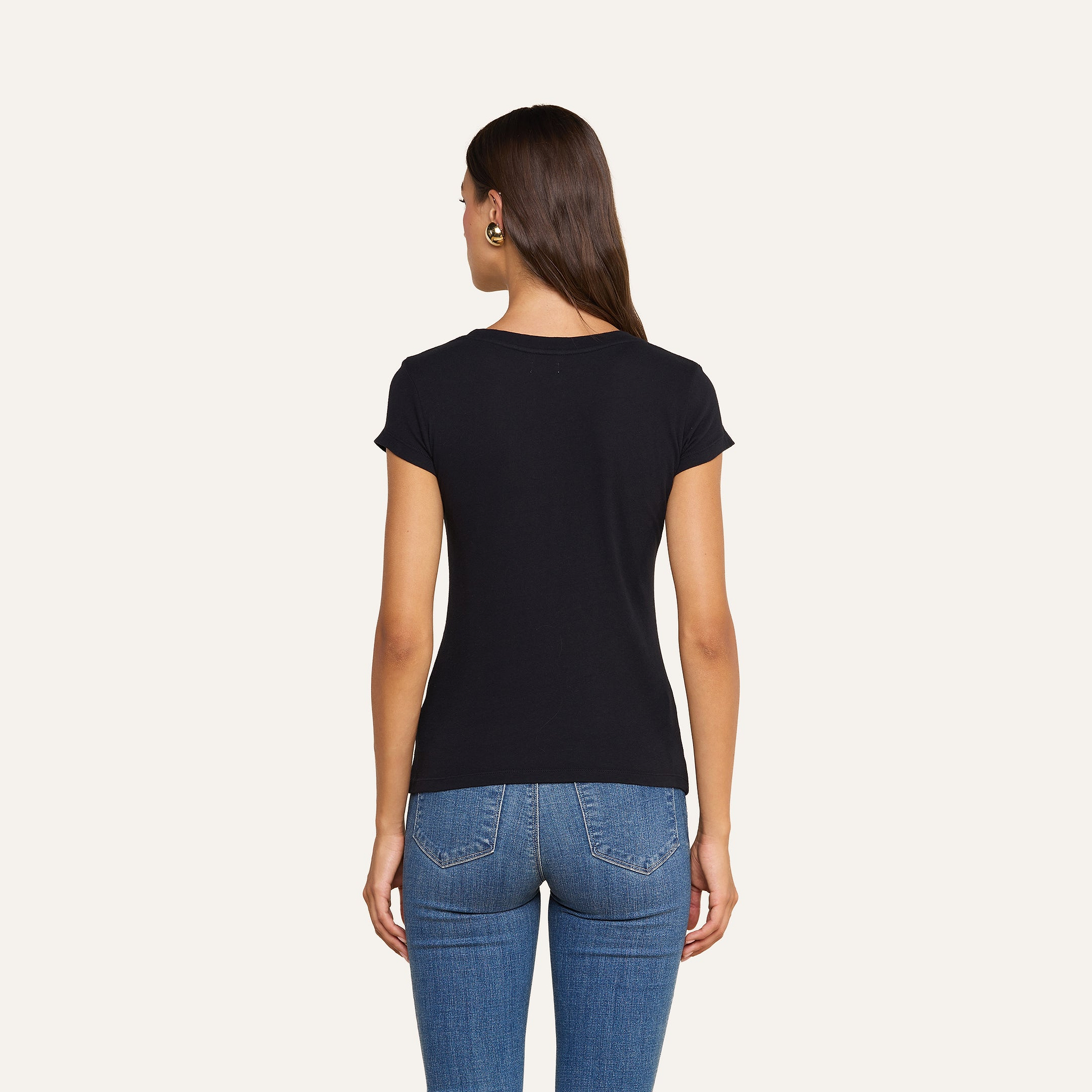Una donna dai lunghi capelli castani, che indossa una maglietta nera a scollo rotondo di Cory Cotton e dei jeans blu, è in piedi di spalle alla fotocamera su uno sfondo beige.