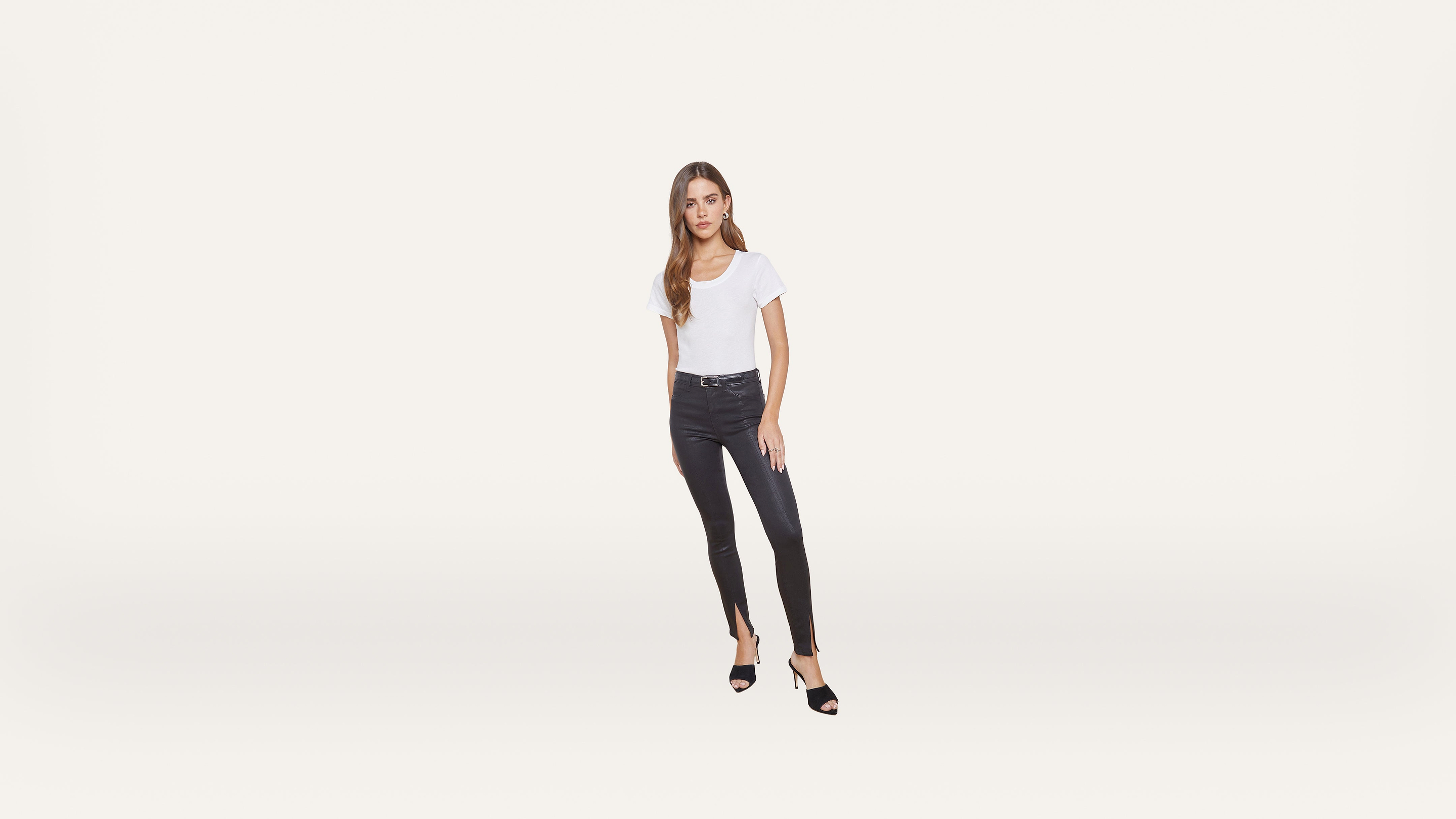 Woman standing against a plain white background, wearing a white t-shirt, black skinny jeans, and black high-heeled sandals. She has long brown hair and a neutral expression, with one hand at her side and the other on her thigh.