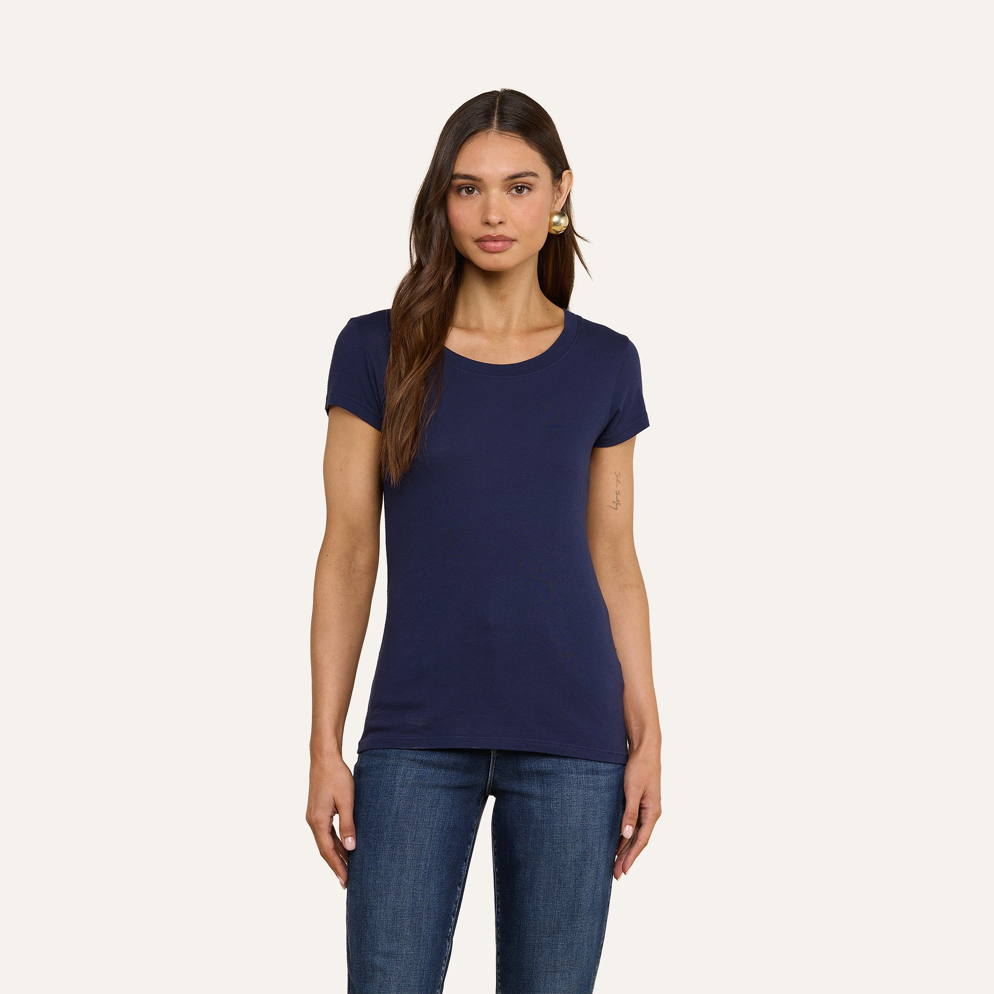 Una donna dai lunghi capelli castani indossa la maglietta con scollo rotondo Cory Cotton e dei jeans blu; è in piedi su uno sfondo beige a tinta unita, con lo sguardo rivolto in avanti e unespressione neutra.
