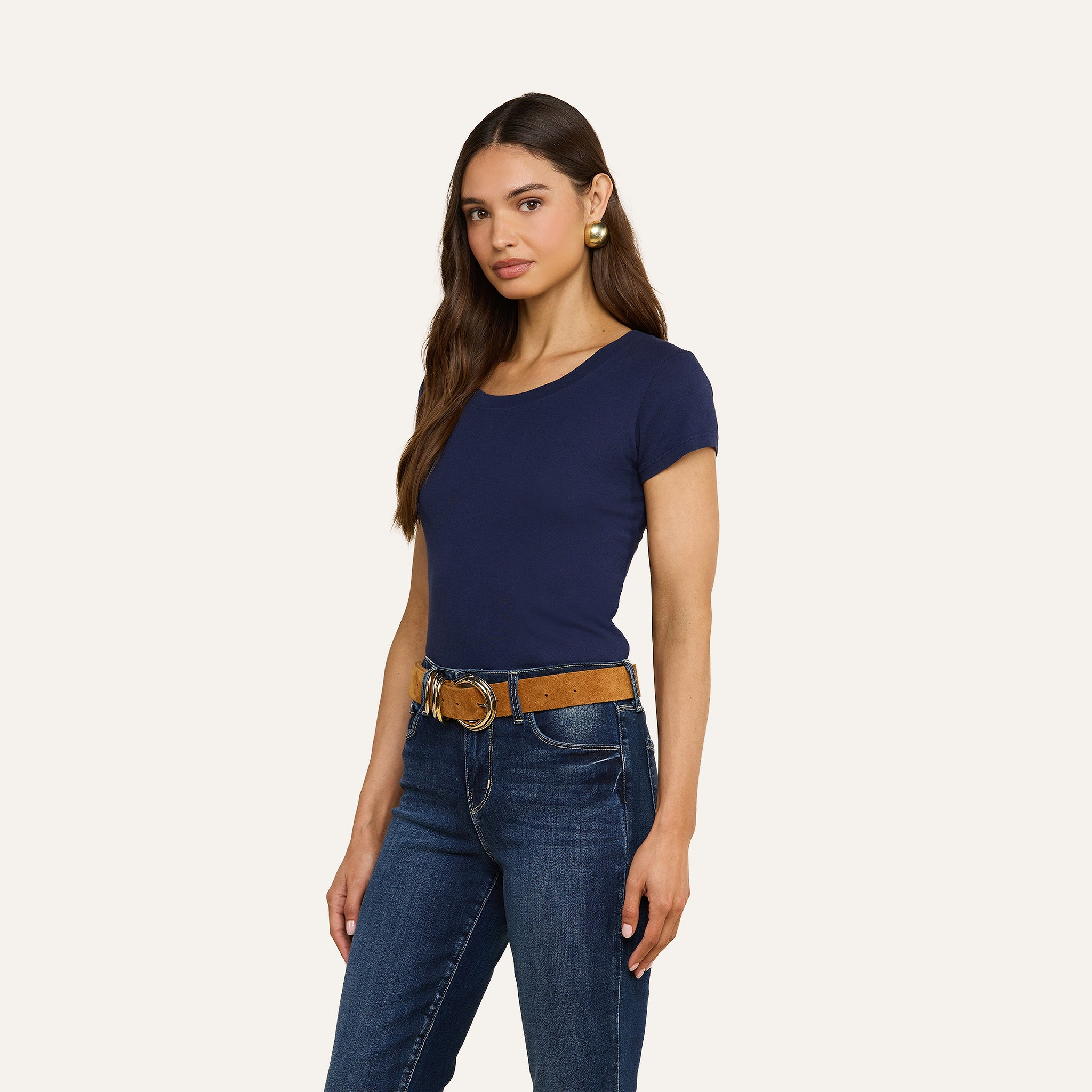 Una donna dai lunghi capelli castani indossa una maglietta blu navy con scollo rotondo della Cory Cotton, jeans blu, una cintura color cuoio e orecchini a cerchio dorati. È in piedi davanti a uno sfondo beige, leggermente girata verso sinistra e con lo sguardo rivolto verso la fotocamera.