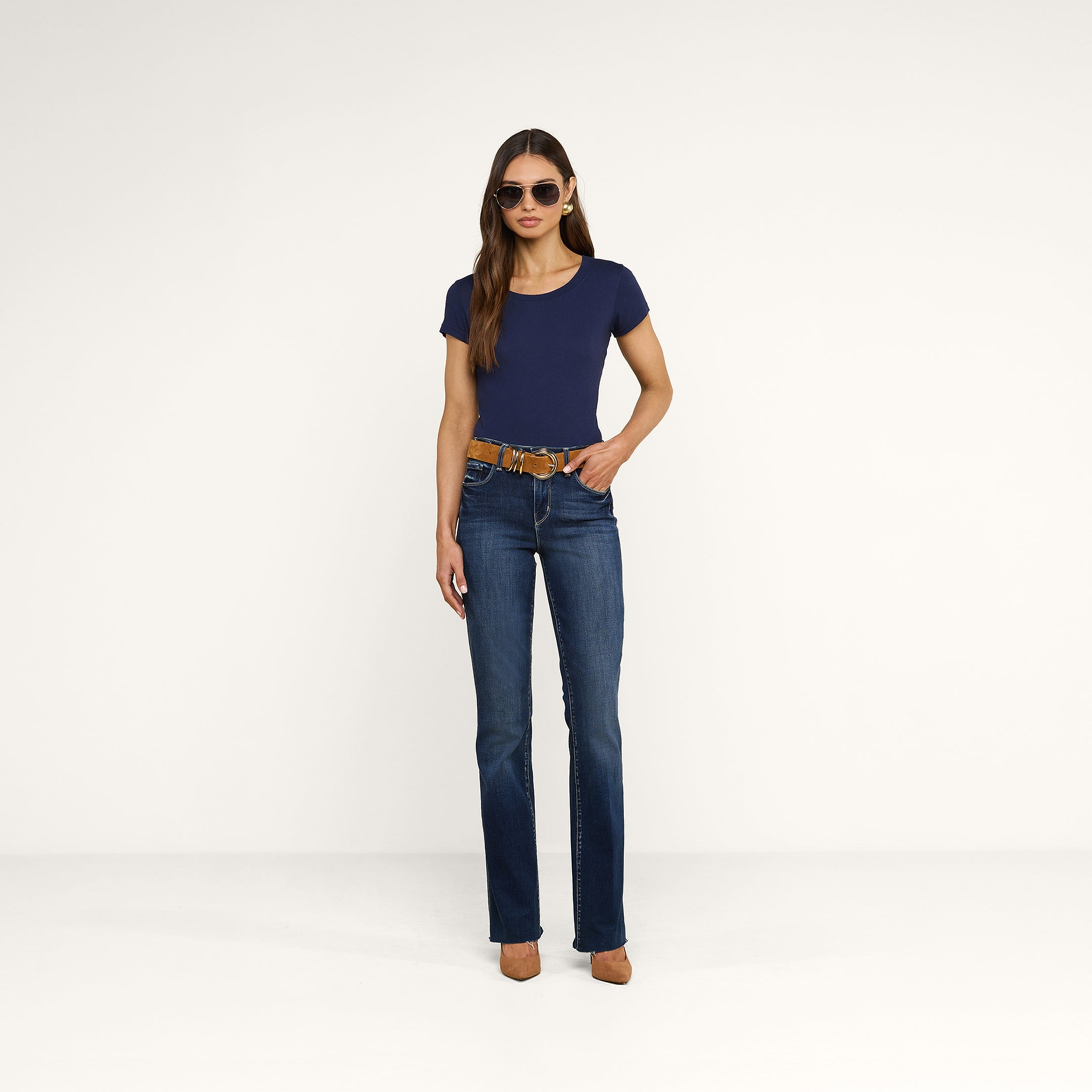 Una donna è in piedi su uno sfondo beige e indossa occhiali da sole, la maglietta Cory Cotton Scoopneck Tee in blu navy, jeans blu con una cintura marrone, tacchi alti color cuoio e ha i capelli lunghi e lisci. Ha le mani appoggiate con disinvoltura nelle tasche anteriori.