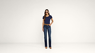 A woman stands against a plain white background, wearing sunglasses, a navy blue t-shirt, blue jeans, a brown belt, and brown high-heeled shoes.