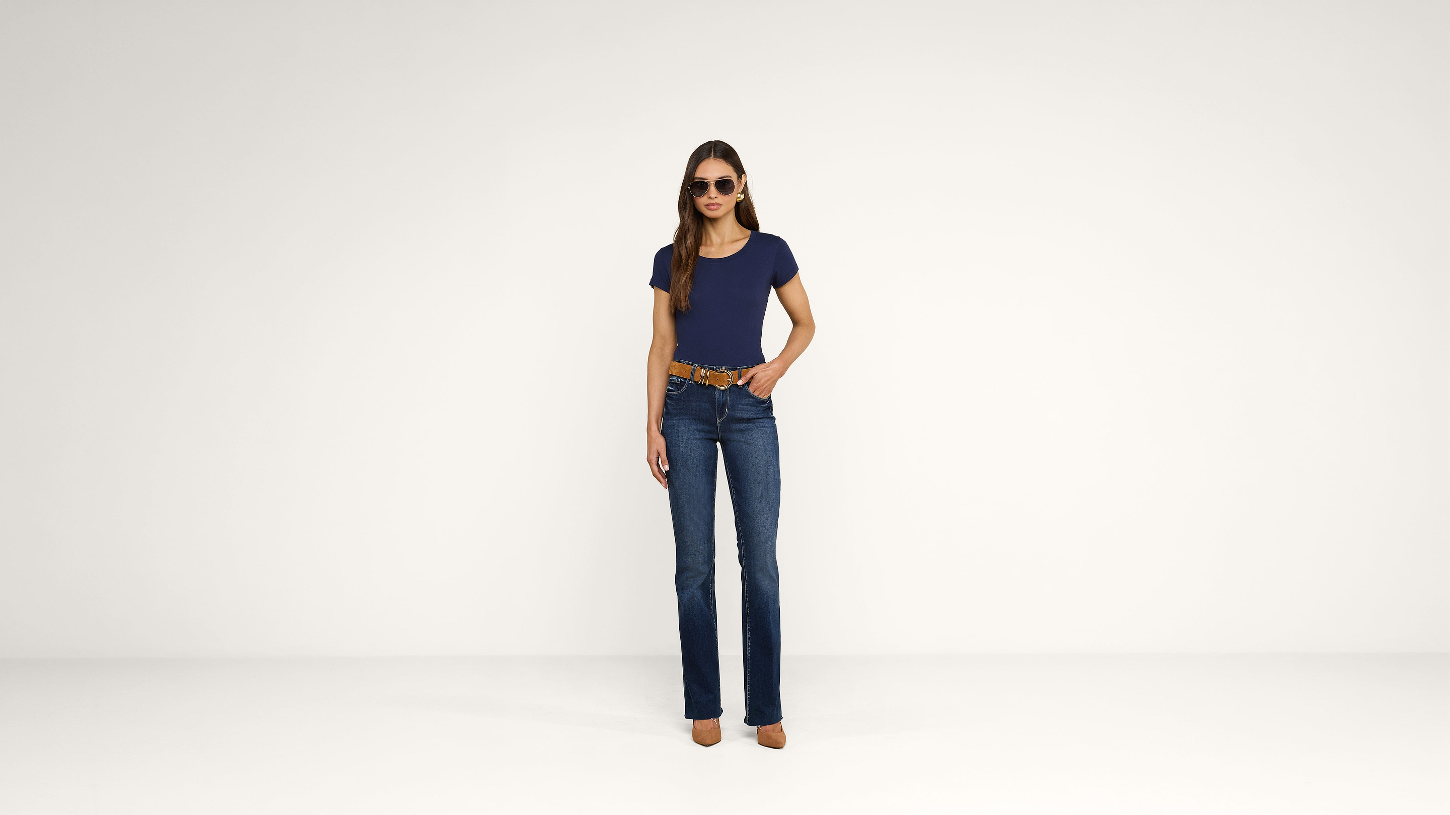 A woman stands against a plain white background, wearing sunglasses, a navy blue t-shirt, blue jeans, a brown belt, and brown high-heeled shoes.