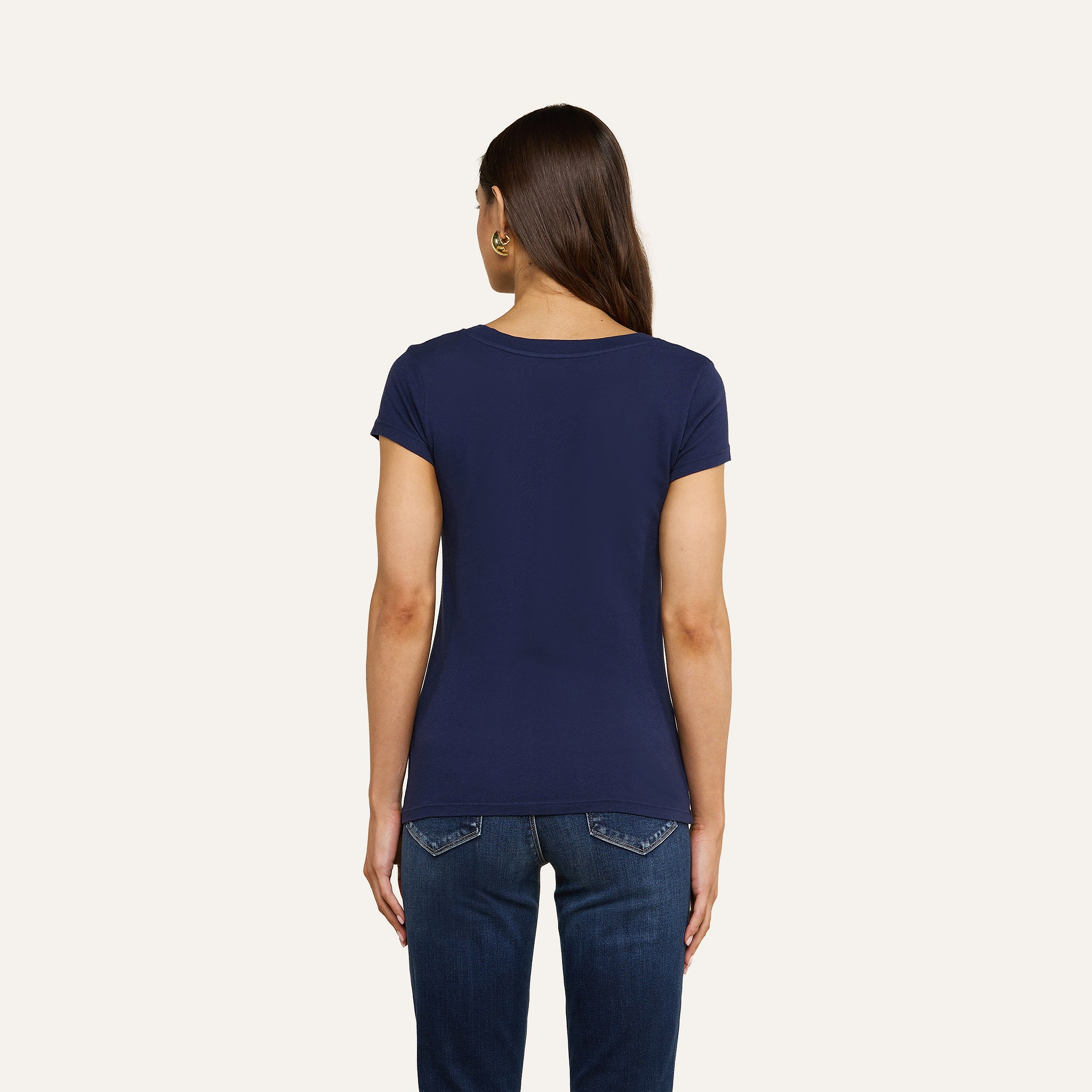 Una donna dai lunghi capelli castani è di spalle; indossa una maglietta blu navy con scollo rotondo di Cory Cotton e jeans blu scuro, su uno sfondo beige a tinta unita.