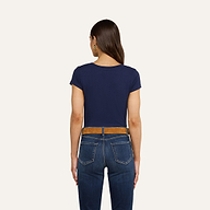 Una donna dai lunghi capelli castani è ritratta di spalle, con indosso una maglietta blu navy a scollo rotondo di Cory Cotton, jeans blu, una cintura marrone e orecchini a cerchio dorati, su uno sfondo beige.
