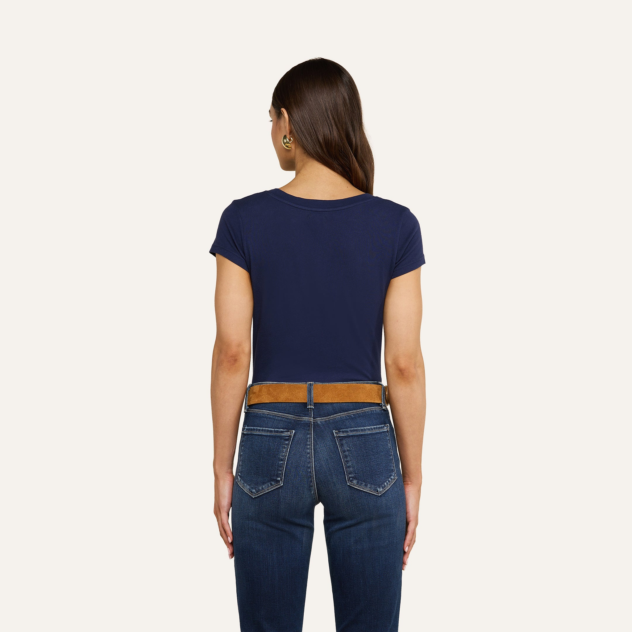Una donna dai lunghi capelli castani è ritratta di spalle, con indosso una maglietta blu navy a scollo rotondo di Cory Cotton, jeans blu, una cintura marrone e orecchini a cerchio dorati, su uno sfondo beige.