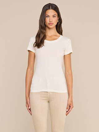 A woman with long brown hair wears the Cory Cotton Scoopneck Tee in vintage off-white and beige pants, standing against a plain beige background, facing the camera with a neutral expression.