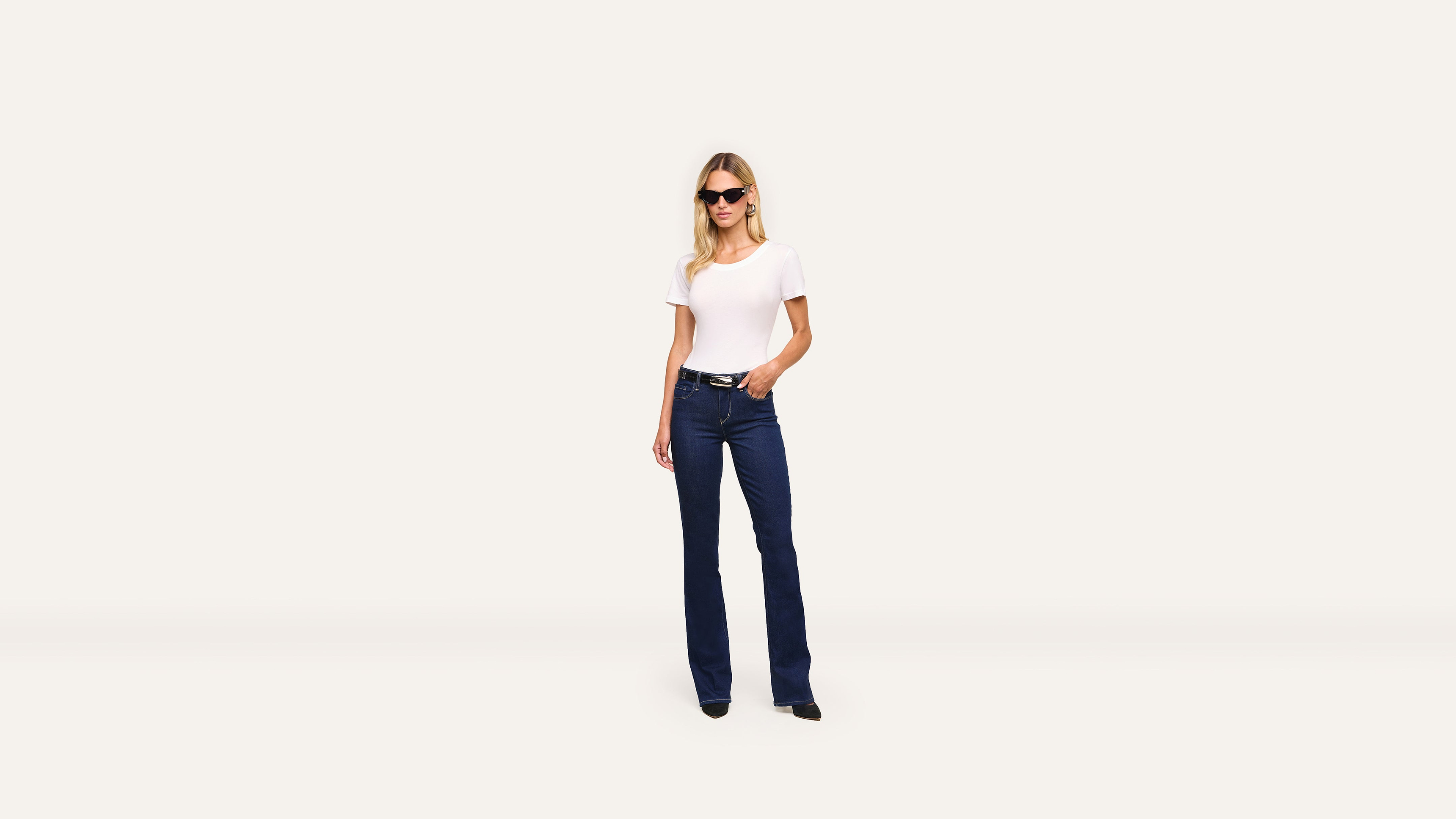 A stylish woman in sunglasses, a white T-shirt, and blue jeans stands confidently against a plain white background.
