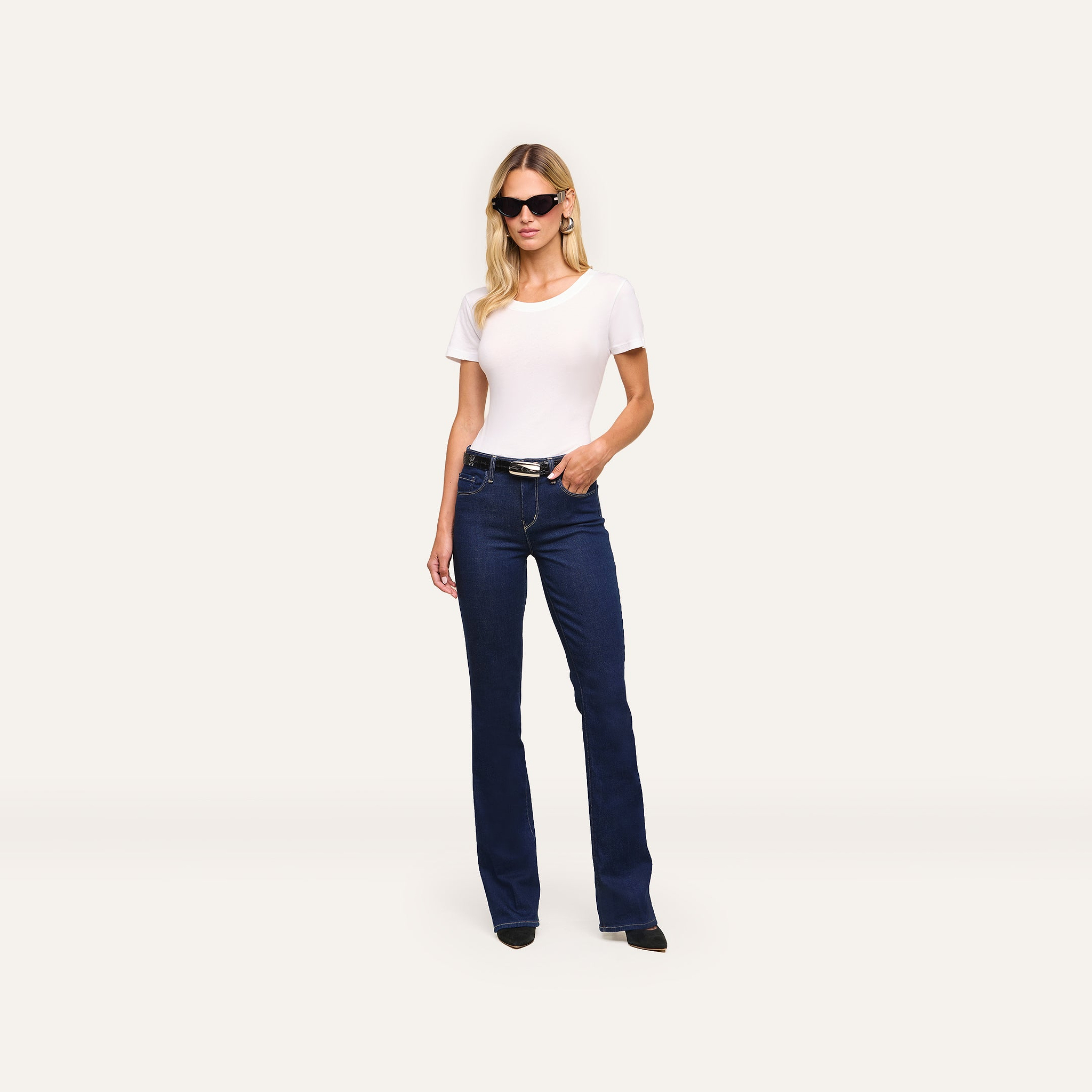 Woman wearing black sunglasses, the Cory Cotton Scoopneck Tee, dark blue bootcut jeans, and black shoes stands against a plain light background with her left hand in her pocket.
