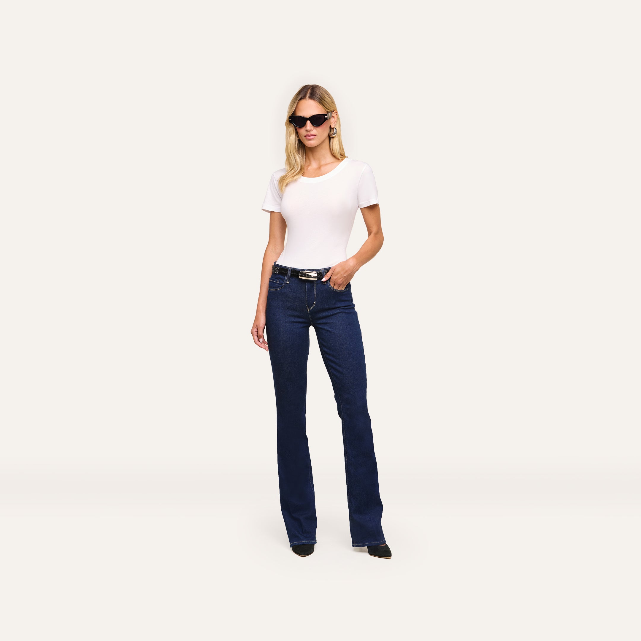 Woman wearing black sunglasses, the Cory Cotton Scoopneck Tee, dark blue bootcut jeans, and black shoes stands against a plain light background with her left hand in her pocket.