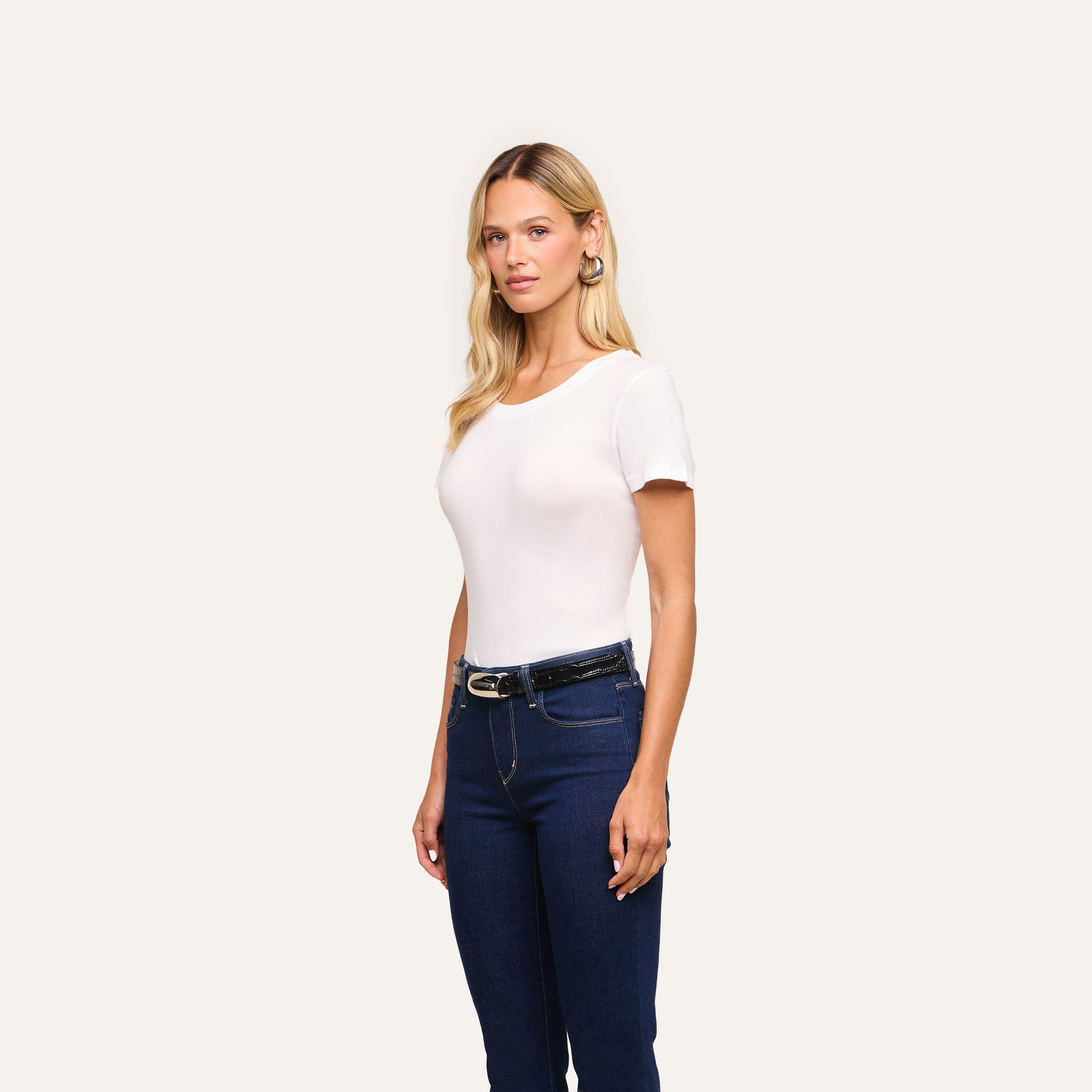A woman with long blonde hair wears the Cory Cotton Scoopneck Tee, blue jeans with a black belt, and hoop earrings, standing against a plain light background.