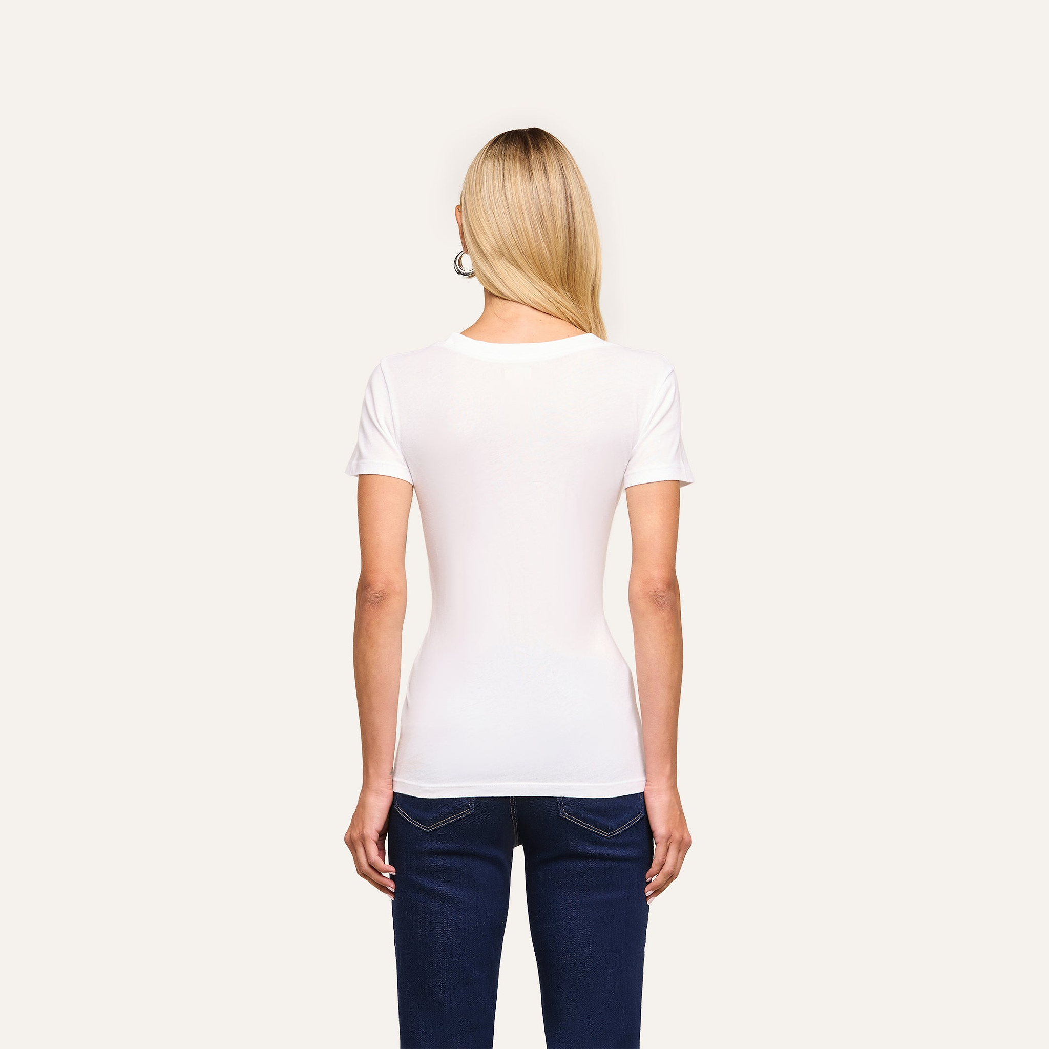 A woman with straight blonde hair, wearing the Cory Cotton Scoopneck Tee in white and dark jeans, stands facing away from the camera against a plain light background.