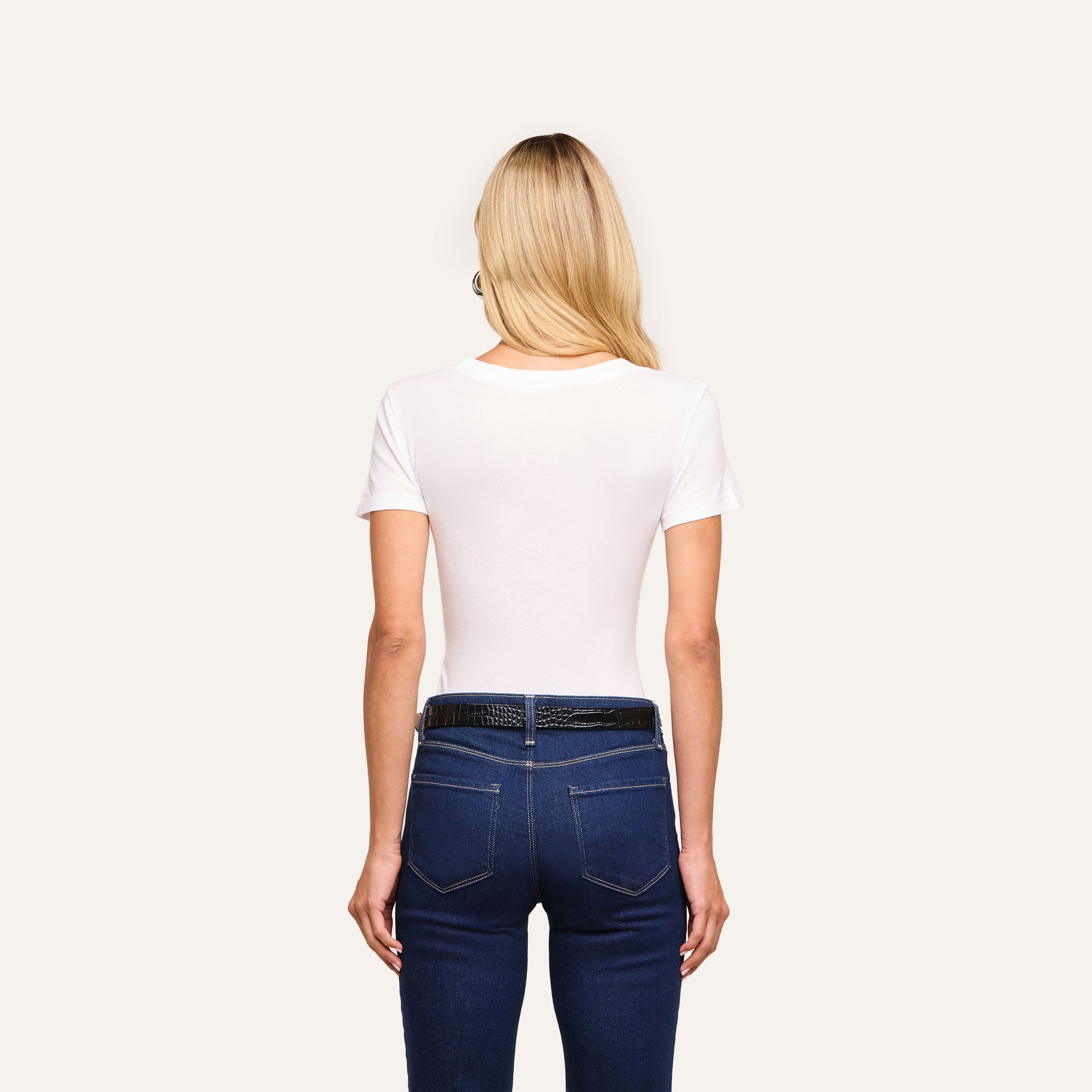 A woman with long blonde hair stands with her back to the camera, wearing the Cory Cotton Scoopneck Tee and dark blue jeans with a black belt against a plain, light background.