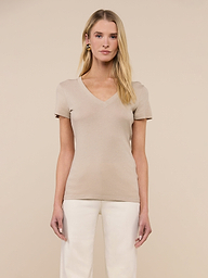 A woman with long blonde hair stands facing forward against a plain beige background, wearing the Becca Cotton V-Neck Tee in a slim fit paired with off-white pants.