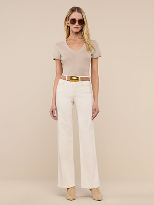 A woman stands in front of a beige background wearing the Becca Cotton V-Neck Tee, high-waisted off-white wide-leg pants, a large belt with a gold buckle, beige boots, sunglasses, and hoop earrings.