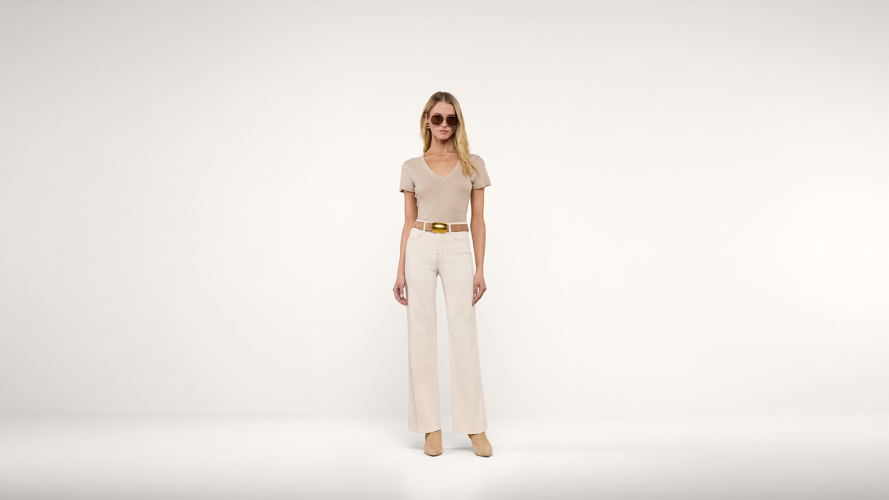 A woman wearing sunglasses, a beige short-sleeve top, wide-leg pants, a tan belt, and matching shoes stands against a plain white background.