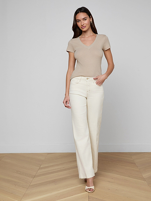 A woman with long brown hair stands indoors on a wooden floor, wearing the slim fit Becca Cotton V-Neck Tee in beige, high-waisted cream wide-leg pants, and heeled sandals. She looks to the side with one hand in her pocket.
