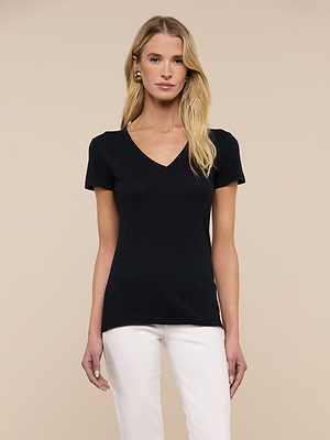 A woman with long blonde hair, wearing the Becca Cotton V-Neck Tee in black and white pants, stands against a plain beige background.