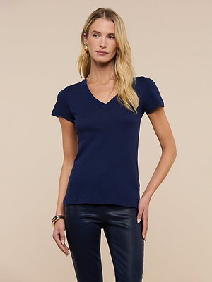 A woman with long blonde hair and a neutral expression wears the Becca Cotton V-Neck Tee in navy, made from 100 cotton, paired with black pants and stands against a plain beige background.