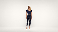 A woman wearing sunglasses, a navy blue top, black pants, a yellow belt, and beige heels stands against a plain white background.
