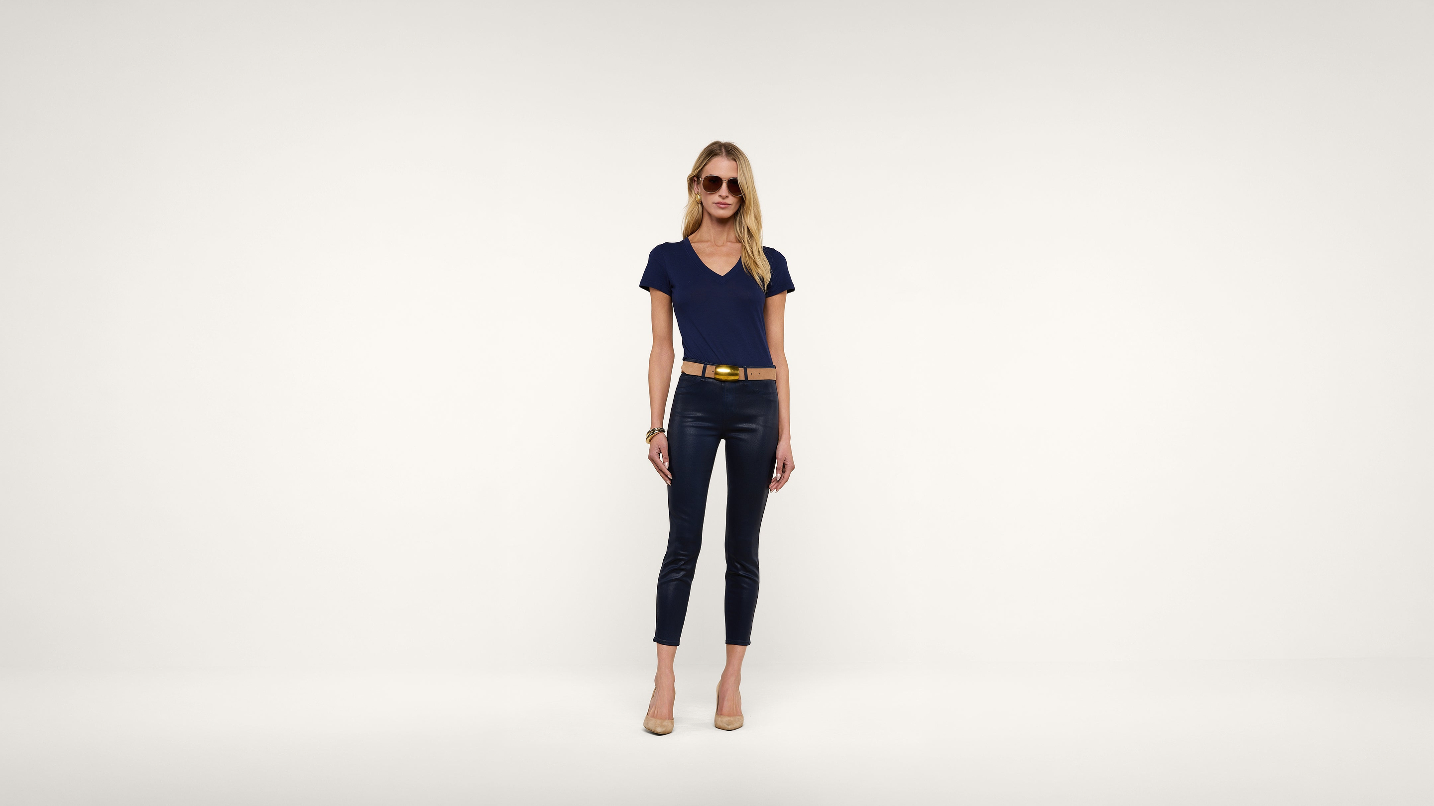 A woman wearing sunglasses, a navy blue top, black pants, a yellow belt, and beige heels stands against a plain white background.