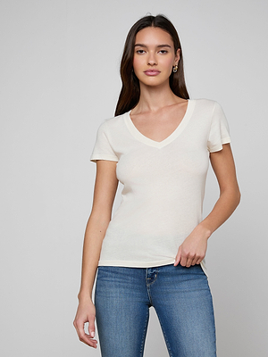 A woman with long brown hair models the Becca Cotton V-Neck Tee, a vintage off-white 100 cotton top, paired with blue jeans. She stands against a light gray background with one hand on her hip and a neutral expression.