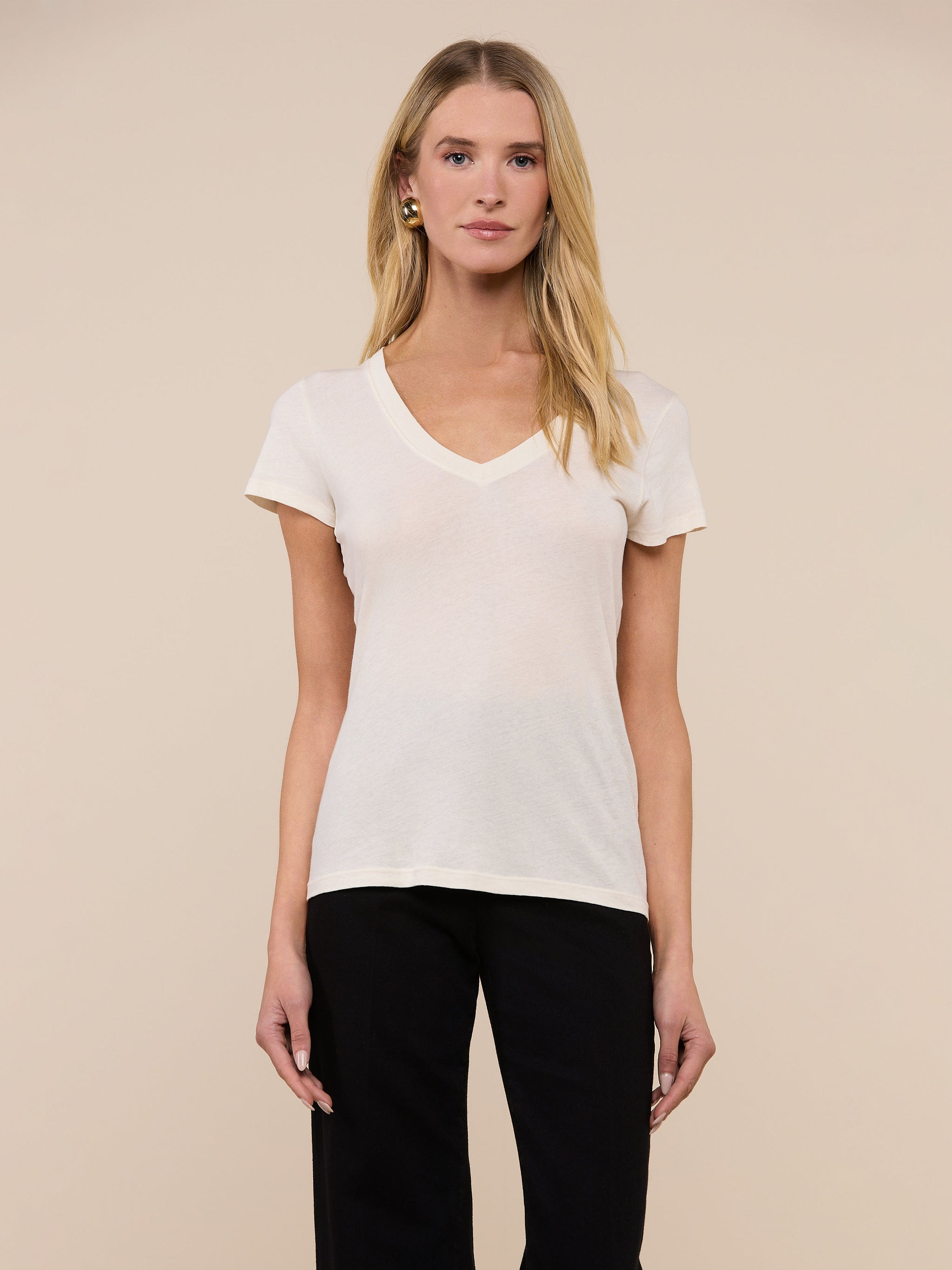 A woman with long blonde hair wears the Becca Cotton V-Neck Tee, an off-white 100 cotton vintage-style top, paired with black pants. She stands against a plain beige background, facing forward with a neutral expression.