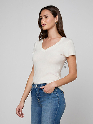 A woman with long brown hair wears the Becca Cotton V-Neck Tee—fitted, vintage off-white, 100 cotton—and blue jeans, standing with one hand in her pocket against a plain light gray background.