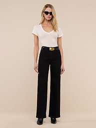 A woman with long brown hair stands indoors on a wooden floor, facing the camera and smiling slightly. She wears the Becca Cotton V-Neck Tee in vintage off-white, blue cropped jeans, and beige heels against a light background.