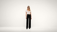 A woman wearing sunglasses, a white top, black wide-leg pants, and black shoes stands against a plain white background.