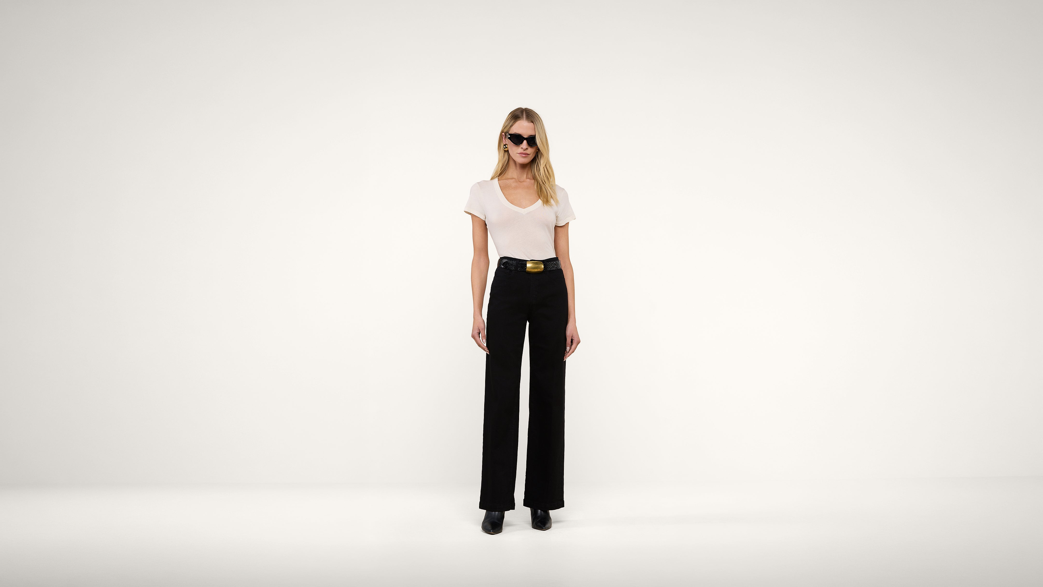 A woman wearing sunglasses, a white top, black wide-leg pants, and black shoes stands against a plain white background.