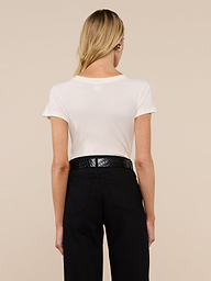 A woman with long brown hair, wearing the Becca Cotton V-Neck Tee in off-white and blue jeans, stands facing away from the camera against a plain light gray background.