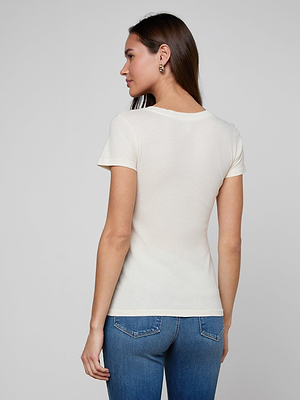 A woman with long brown hair, wearing the Becca Cotton V-Neck Tee in off-white and blue jeans, stands facing away from the camera against a plain light gray background.
