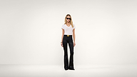 Woman wearing sunglasses, a white t-shirt, black flared pants, and a black belt stands against a plain white background.