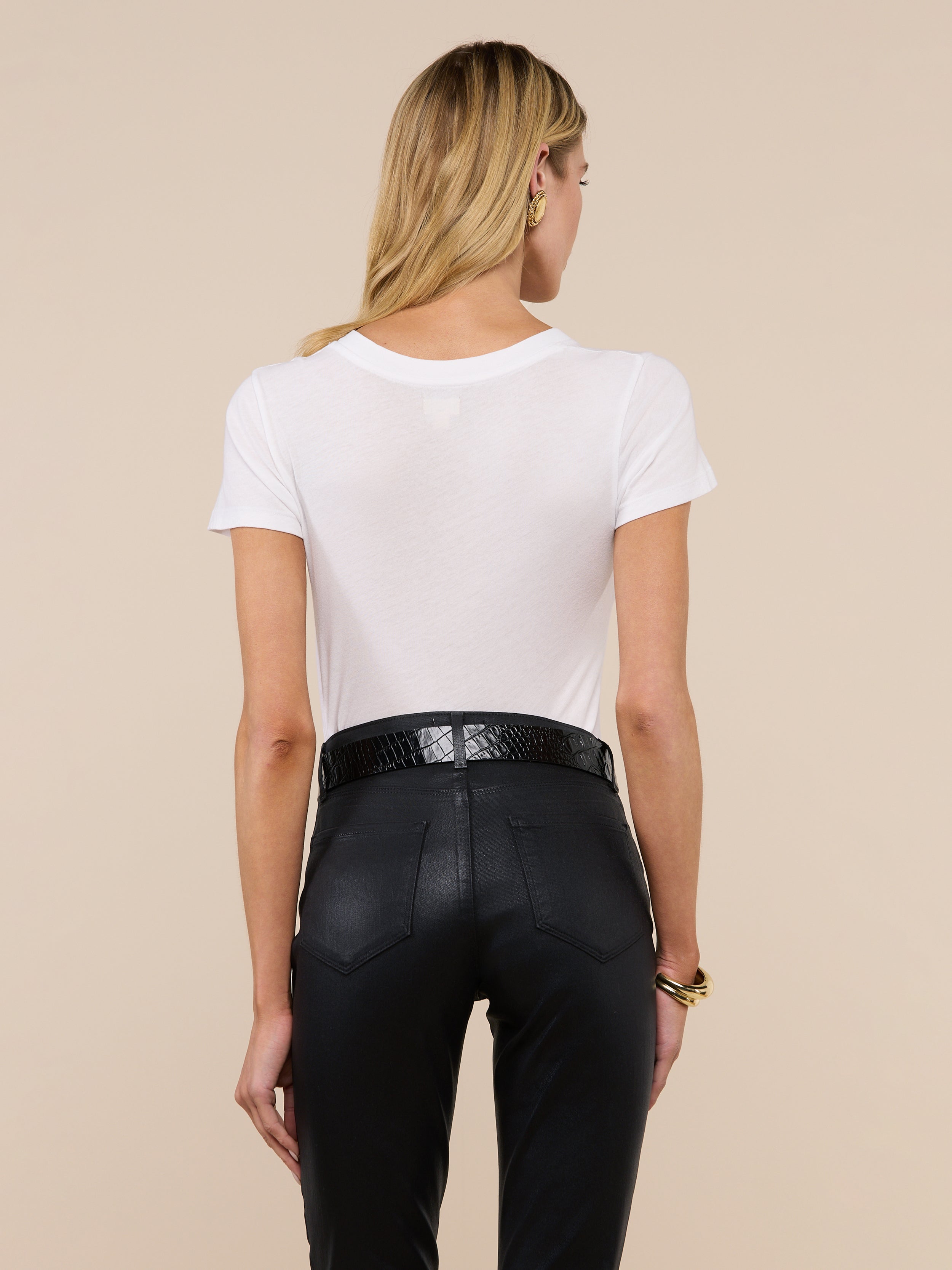 A woman with long blonde hair is seen from behind, wearing the Becca Cotton V-Neck Tee in white, tucked into high-waisted black pants with a black belt, against an off-white background.