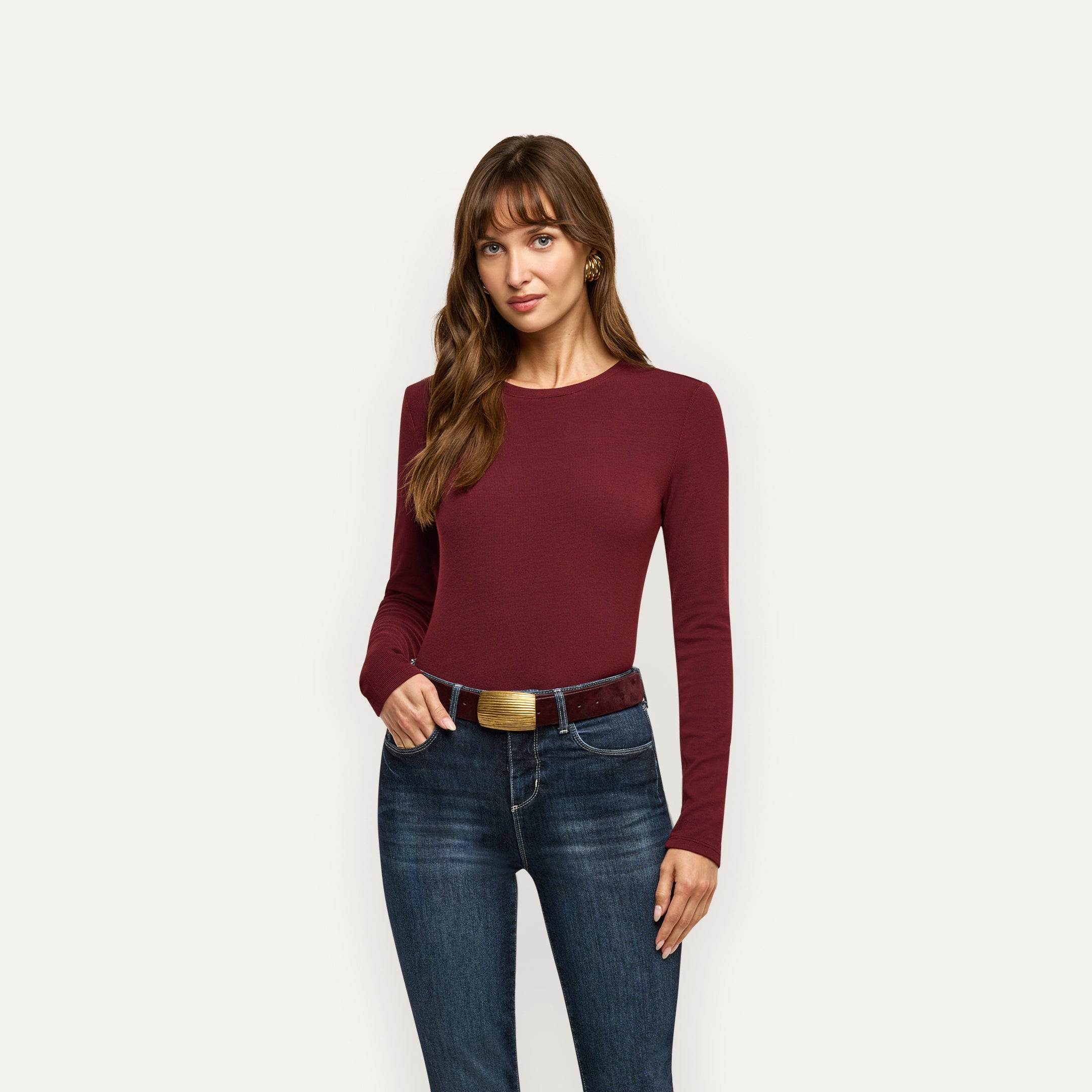 A woman wearing a slim-fit, long-sleeved burgundy Tess Long Sleeve Tee tucked into dark wash jeans with a brown belt.