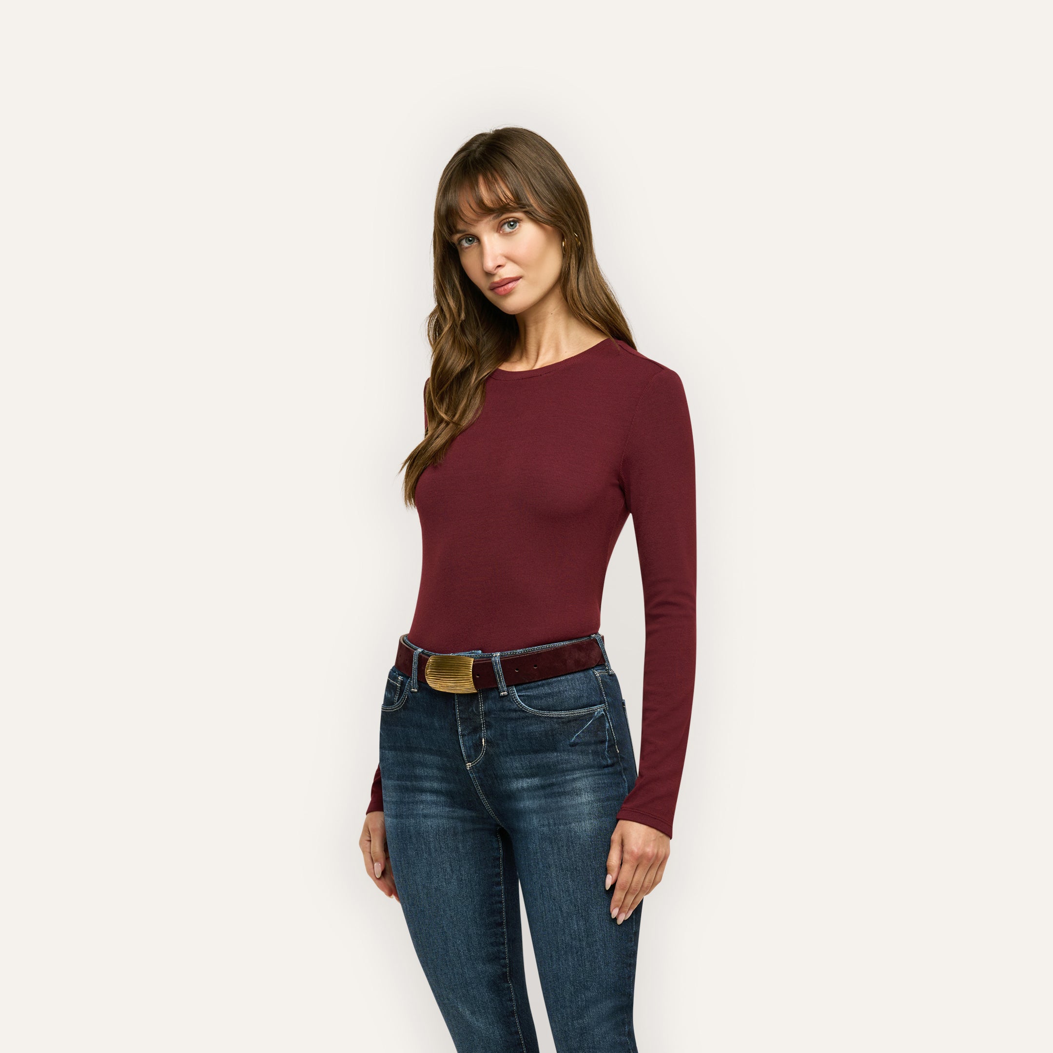A person wearing a slim-fit, long-sleeved burgundy Tess Long Sleeve Tee tucked into dark wash jeans with a belt.