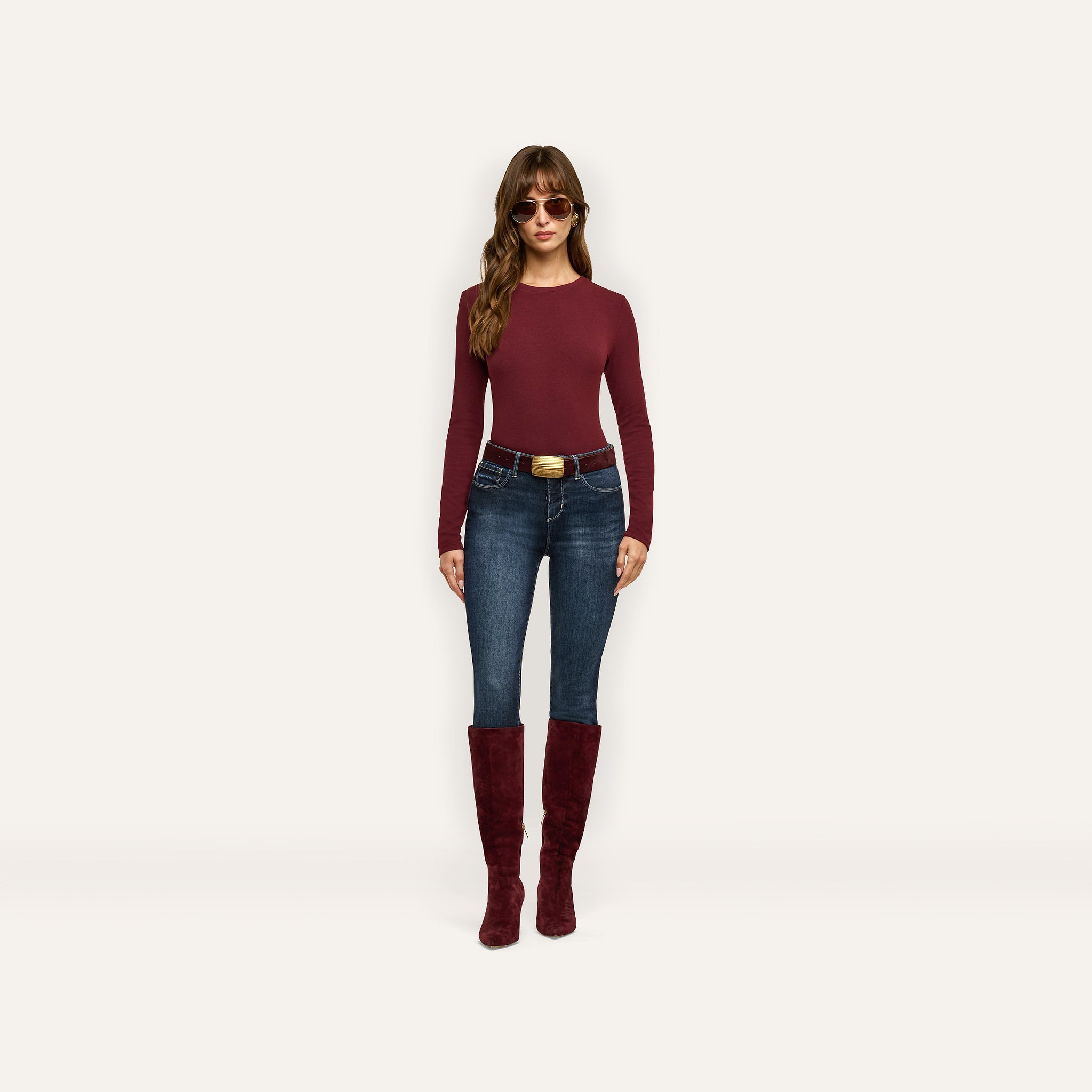 A woman wearing a maroon Tess Long Sleeve Tee, dark wash jeans with a large gold-buckled belt, and knee-high maroon suede boots.