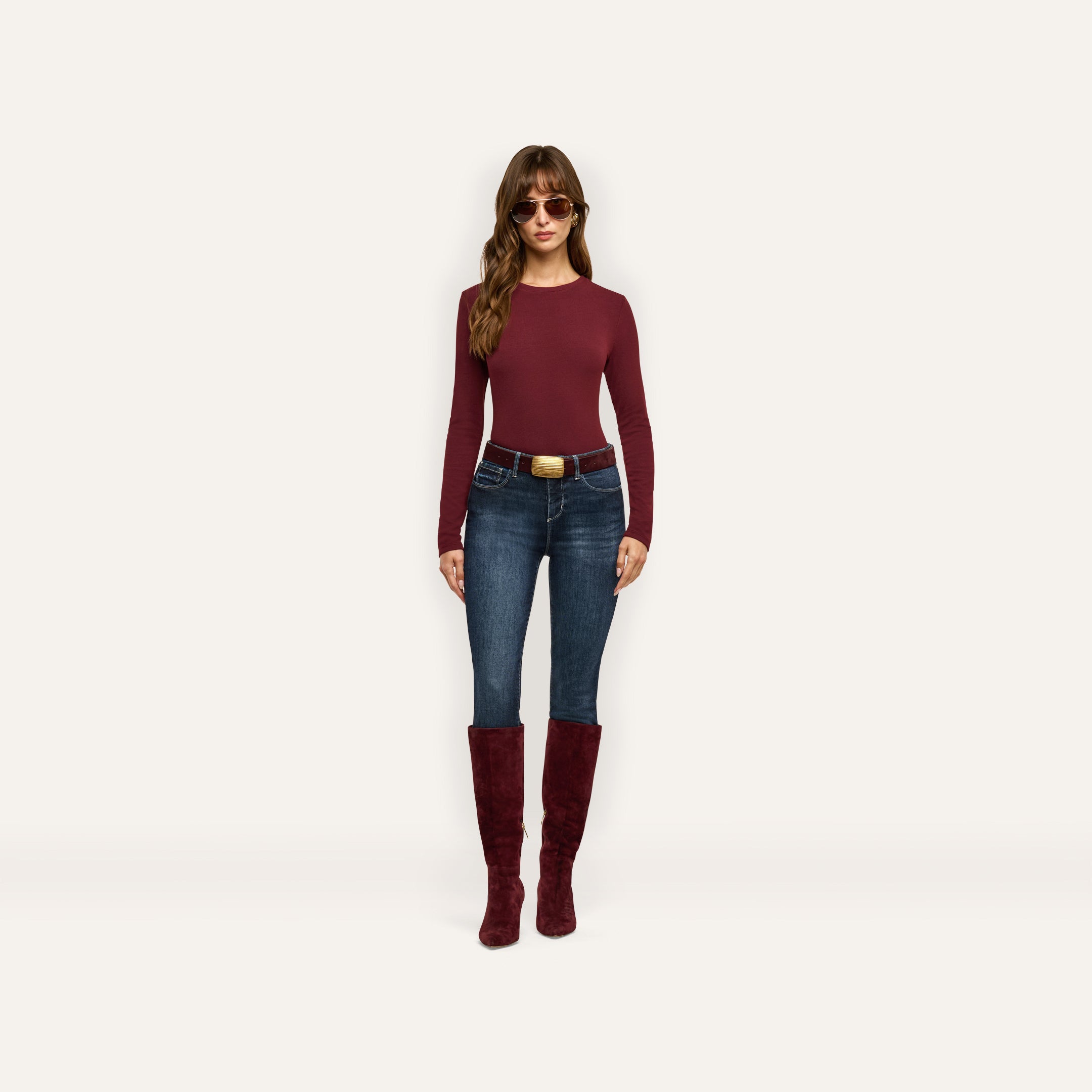 A woman wearing a maroon Tess Long Sleeve Tee, dark wash jeans with a large gold-buckled belt, and knee-high maroon suede boots.