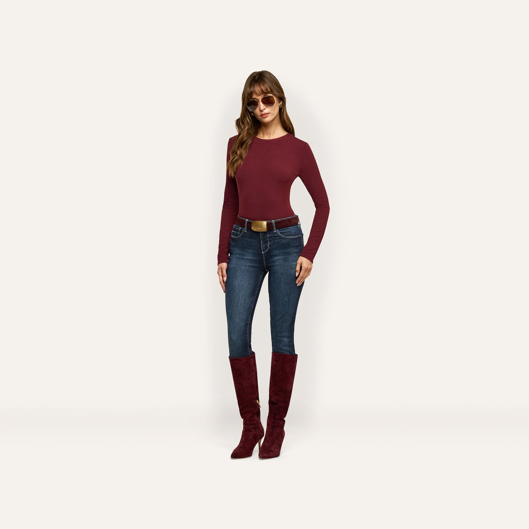 A woman wearing a maroon Tess Long Sleeve Tee, dark-wash jeans with a brown belt, and matching maroon knee-high boots, accessorized with sunglasses.