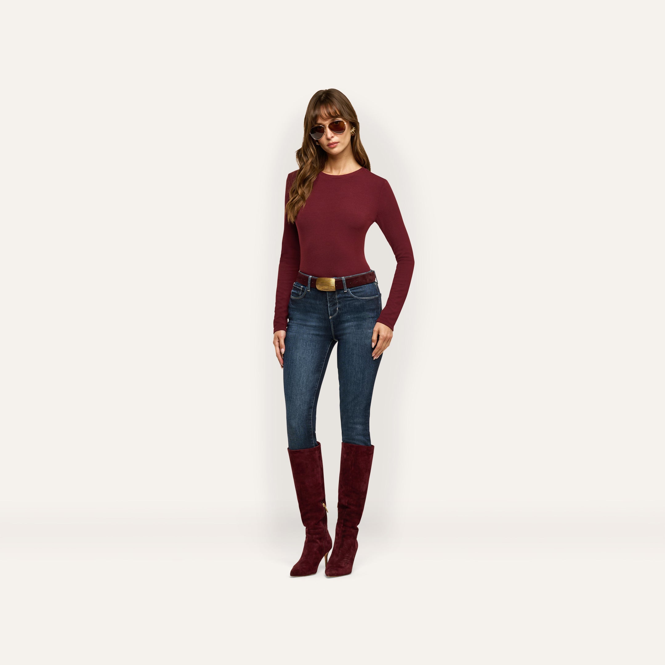 A woman wearing a maroon Tess Long Sleeve Tee, dark-wash jeans with a brown belt, and matching maroon knee-high boots, accessorized with sunglasses.