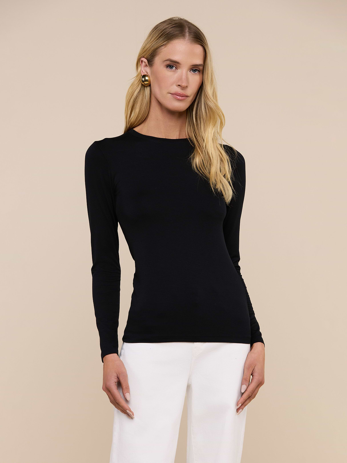 A woman with long blonde hair wears the Tess Long Sleeve Tee in black and white pants, standing against a plain beige background and looking at the camera with a neutral expression.
