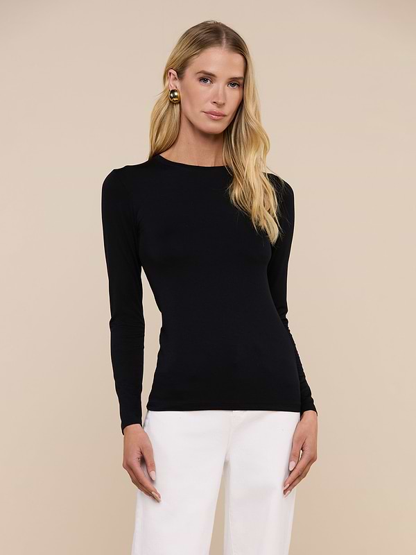 A person with long blonde hair wears the fitted black Tess Long Sleeve Tee and white pants, standing against a plain light background.