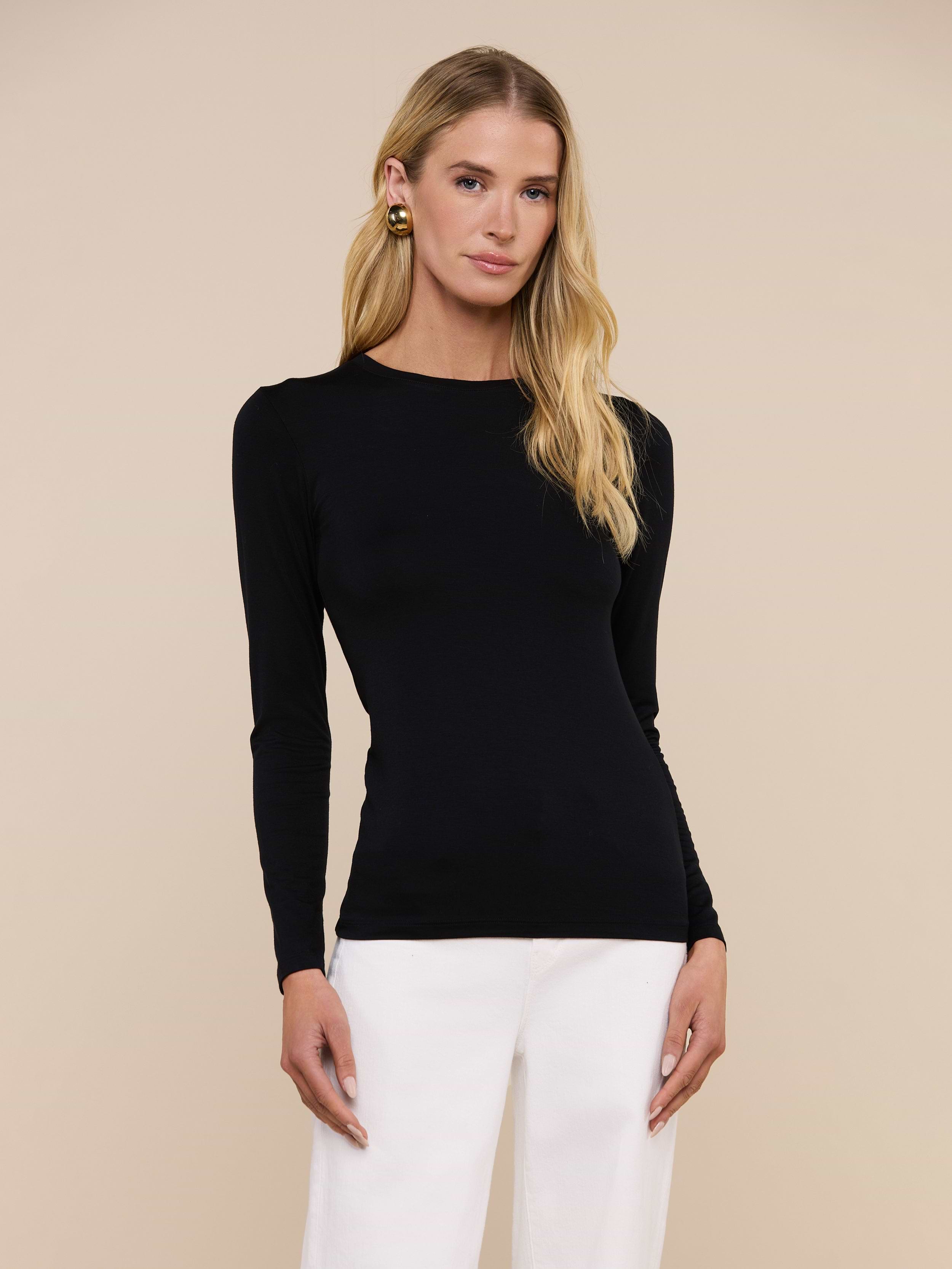 A person with long blonde hair wears the fitted black Tess Long Sleeve Tee and white pants, standing against a plain light background.