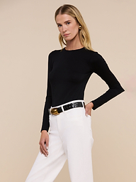 A woman stands confidently against a plain light background, wearing the Tess Long Sleeve Tee in black with white high-waisted pants and a black belt, looking directly at the camera.