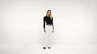 A woman stands against a plain white background, wearing a black long-sleeve top, white wide-leg pants, a black belt, and black shoes. She has long blonde hair and a neutral expression.