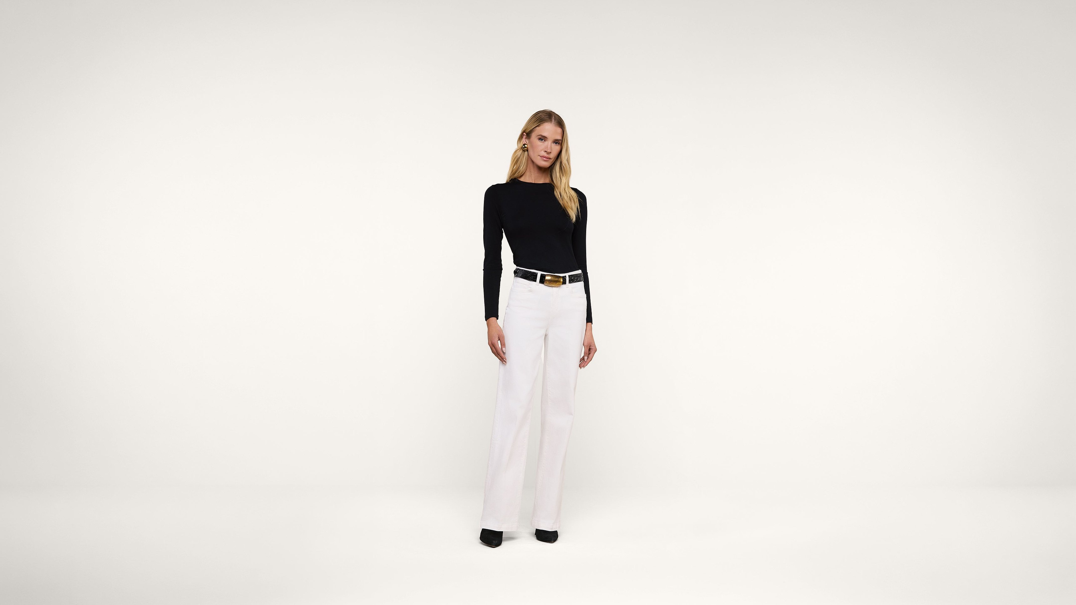 A woman stands against a plain white background, wearing a black long-sleeve top, white wide-leg pants, a black belt, and black shoes. She has long blonde hair and a neutral expression.