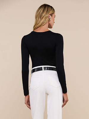 A woman with long blonde hair stands facing away, wearing the fitted black Tess Long Sleeve Tee, white high-waisted pants, a black textured belt, and black earrings against a plain beige background.